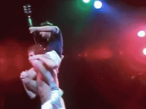 A Man Is Carrying Another Man On His Shoulders On A Stage While Holding A Guitar . GIF