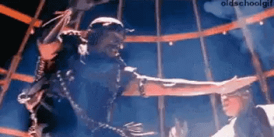 A Man Is Chained To A Pole And Reaching Out Towards Another Man . GIF