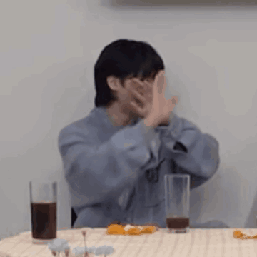 A Man Is Covering His Face With His Hands While Sitting At A Table With A Glass Of Soda . GIF