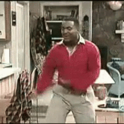 A Man Is Dancing In A Living Room Wearing A Red Sweater And Khaki Pants . GIF