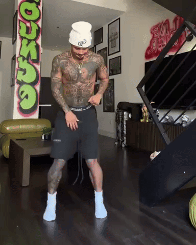 A Man Is Dancing In A Living Room With A Graffiti Wall That Says ' Dunzo ' On It GIF