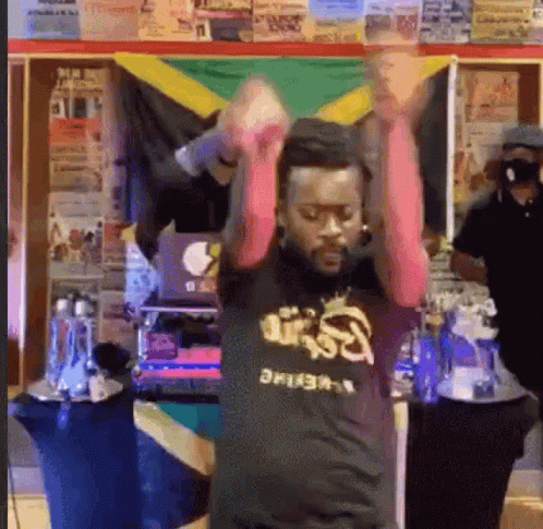 A Man Is Dancing In Front Of A Jamaican Flag While Wearing A Black Shirt . GIF