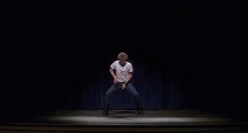 A Man Is Dancing On A Stage In A Dark Room With A Blue Curtain Behind Him . GIF