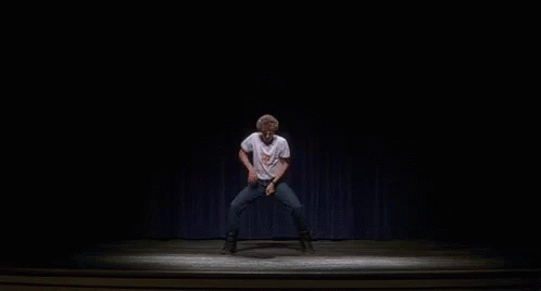 A Man Is Dancing On A Stage In A Dark Room With A Blue Curtain Behind Him . GIF