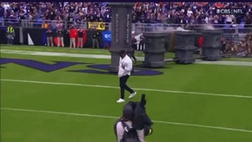 A Man Is Doing A Trick On A Football Field In Front Of A Ravens Sign GIF