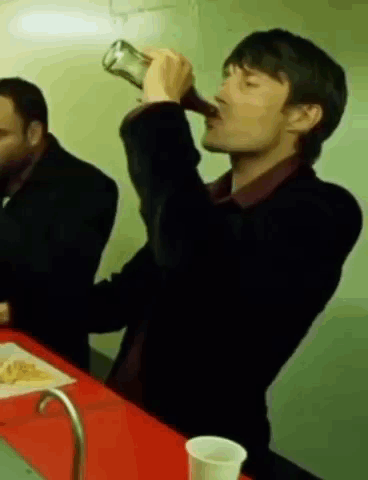 A Man Is Drinking From A Bottle While Sitting At A Table With Another Man . GIF