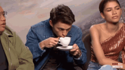 A Man Is Drinking From A Cup While A Woman Sits Behind Him GIF