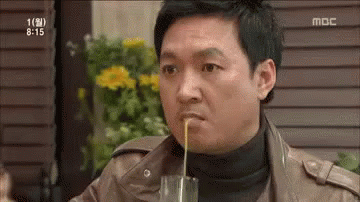 A Man Is Drinking Something Through A Straw From A Glass While Sitting At A Table . GIF