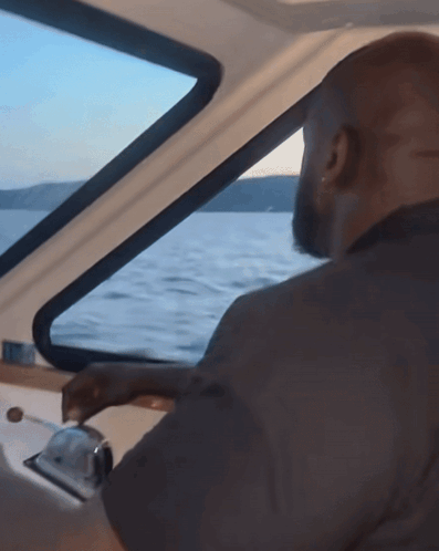 A Man Is Driving A Boat In The Water And Looking Out The Window GIF