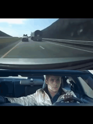 A Man Is Driving A Car On A Highway With A Truck Behind Him GIF