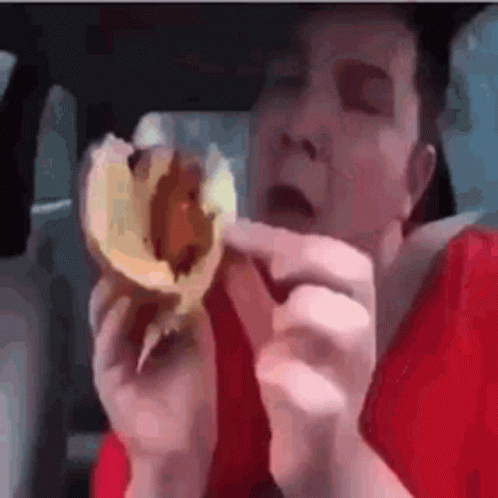 A Man Is Eating A Hamburger In A Car While Wearing A Red Shirt . GIF