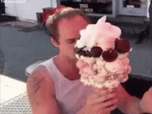 A Man Is Eating A Large Ice Cream Cone With Whipped Cream And Cherries On It . GIF