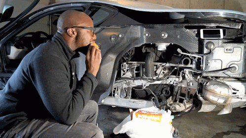 A Man Is Eating A Sandwich In Front Of A Car That Is Being Built GIF