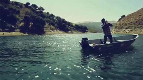 A Man Is Fishing In A Boat On A Lake With Mountains In The Background . GIF
