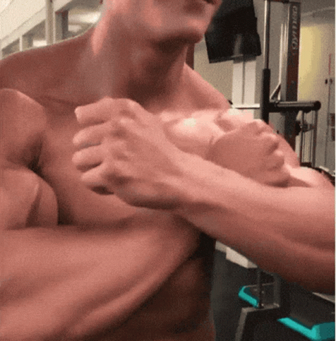 A Man Is Flexing His Muscles In A Gym While Holding His Arms Together . GIF