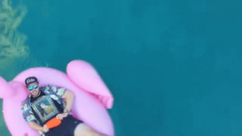 A Man Is Floating On A Pink Flamingo Float In The Water GIF
