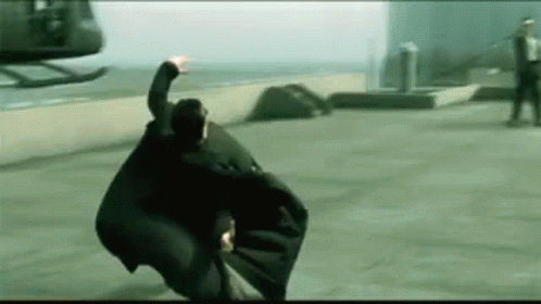 A Man Is Flying Through The Air In Front Of A Helicopter While Wearing A Black Cape . GIF