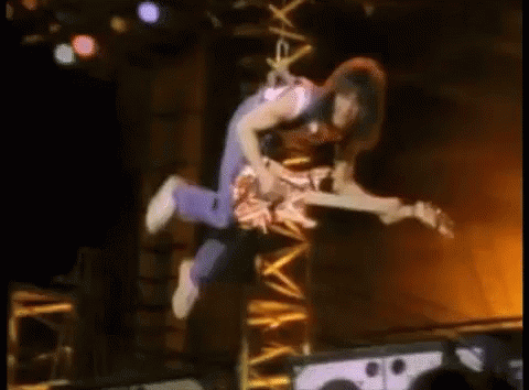 A Man Is Flying Through The Air While Holding A Guitar . GIF