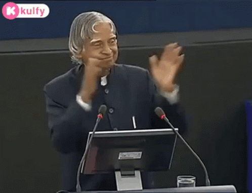 A Man Is Giving A Speech In Front Of A Microphone And A Computer Monitor . GIF