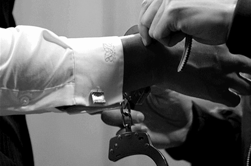 A Man Is Handcuffed To Another Man 'S Arm With A White Shirt With The Letter B On The Sleeve GIF