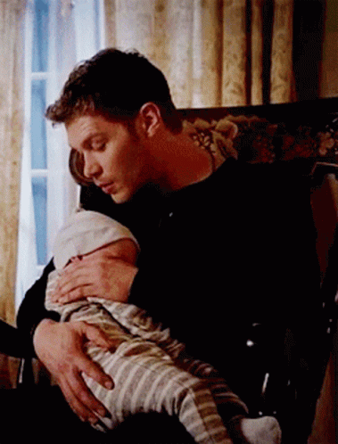 A Man Is Holding A Baby In His Lap While Sitting In A Chair . GIF