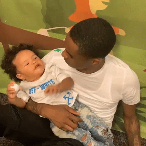 A Man Is Holding A Baby Who Is Wearing A White Shirt That Says Off White GIF