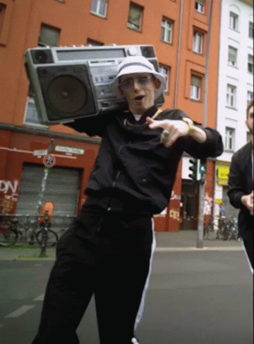 A Man Is Holding A Boombox Over His Head In Front Of A Building That Says Dn GIF