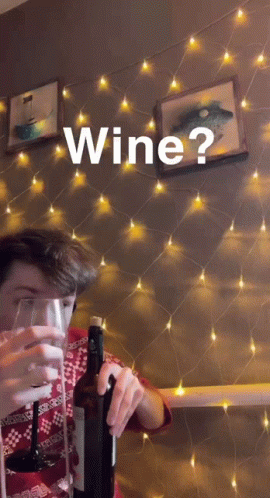 A Man Is Holding A Bottle Of Wine And A Glass In Front Of A String Of Lights And The Word Wine Is On The Wall GIF