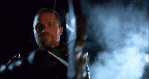 A Man Is Holding A Bow And Arrow In Front Of A Smokey Background . GIF