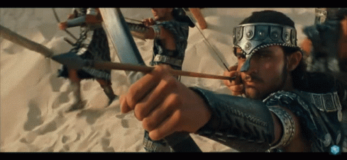 A Man Is Holding A Bow And Arrow With A Few Other Men In The Background GIF