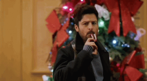 A Man Is Holding A Candy Cane In His Mouth While Standing In Front Of A Christmas Tree . GIF