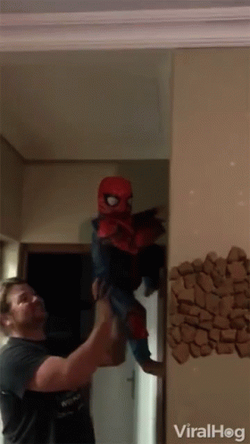 A Man Is Holding A Child In A Spiderman Costume In Front Of A Wall That Says Viralhog GIF