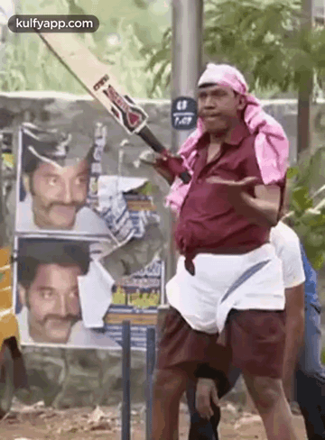 A Man Is Holding A Cricket Bat In Front Of A Wall With Posters Of Actors On It . GIF