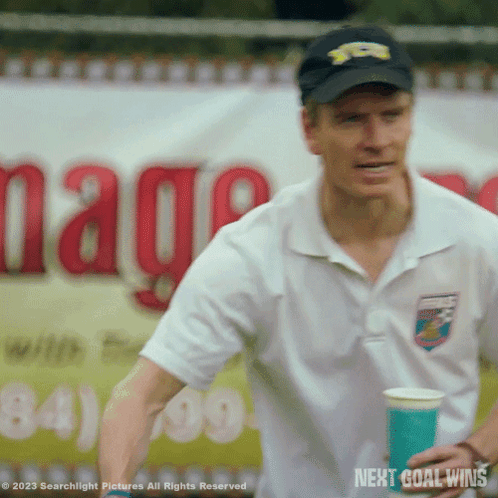 A Man Is Holding A Cup In Front Of A Sign That Says " Next Goal Wins " GIF
