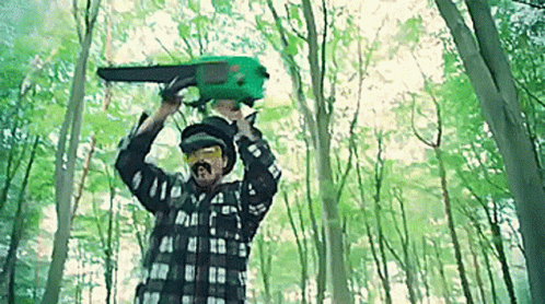 A Man Is Holding A Green Chainsaw Over His Head In The Woods . GIF