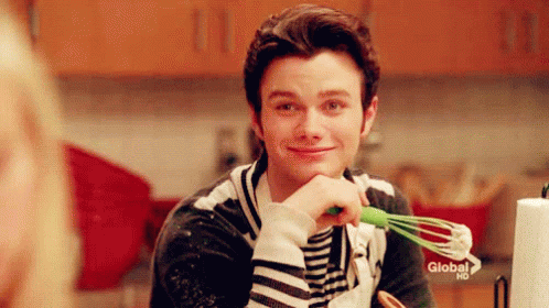 A Man Is Holding A Green Whisk In His Hand And Smiling In A Kitchen . GIF
