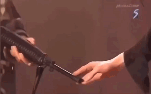 A Man Is Holding A Gun In Front Of A Man Standing Next To A Man Holding A Gun . GIF