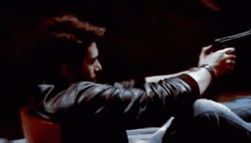 A Man Is Holding A Gun In His Hand While Sitting In A Chair . GIF