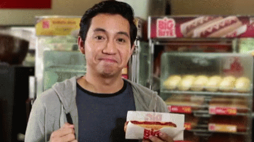 A Man Is Holding A Hot Dog In Front Of A Big Hot Dog Stand GIF