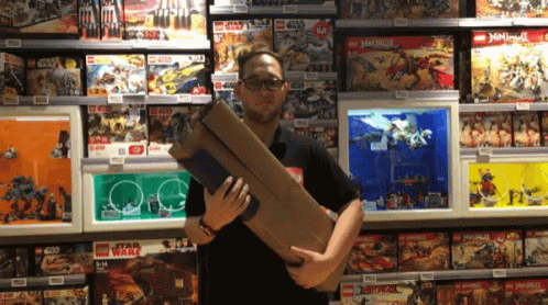 A Man Is Holding A Large Box In Front Of A Display Of Lego Star Wars And Ninjago Toys GIF