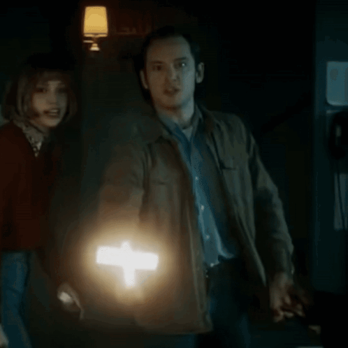 A Man Is Holding A Lighted Cross In Front Of A Woman In A Dark Room . GIF