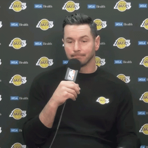 A Man Is Holding A Microphone In Front Of A Wall With Lakers Logos GIF