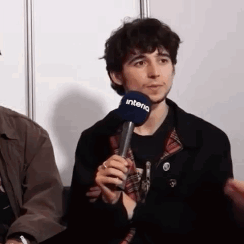 A Man Is Holding A Microphone In His Hand While Sitting In Front Of A Wall . GIF