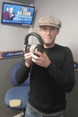 A Man Is Holding A Pair Of Headphones In Front Of A Big Show Sign GIF