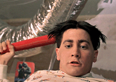 A Man Is Holding A Red Object In Front Of His Head With A Picture Of A Man On The Wall In The Background GIF