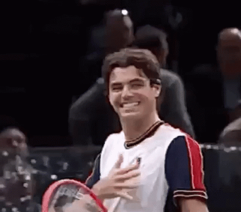 A Man Is Holding A Tennis Racquet And Smiling While Standing On A Tennis Court . GIF
