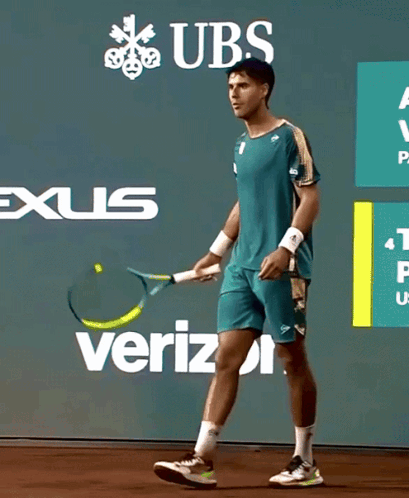 A Man Is Holding A Tennis Racquet In Front Of A Wall With Ubs And Verizon Advertisements GIF
