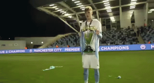 A Man Is Holding A Trophy On A Soccer Field With Manchester City Premier League Banners Behind Him GIF