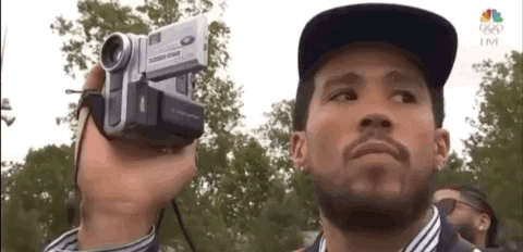 A Man Is Holding A Video Camera In His Hand And Looking At The Camera . GIF