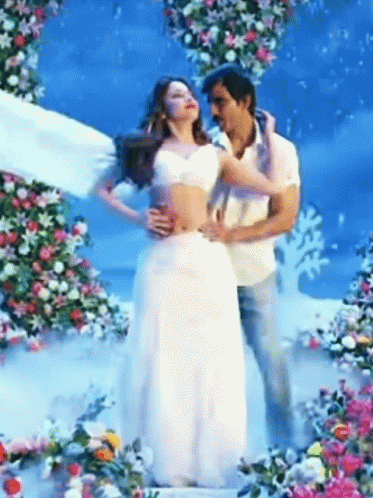 A Man Is Holding A Woman In A White Dress In Front Of Flowers GIF